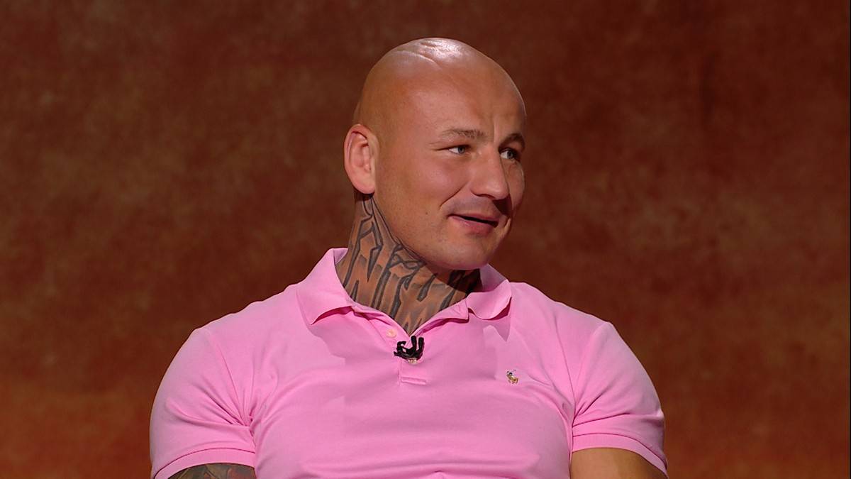 Mężczyzna z ogoloną głową i tatuażami na szyi, ubrany w r&oacute;żową koszulkę polo, siedzi i uśmiecha się.