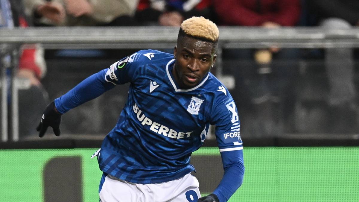 Dwóch piłkarzy grających w piłkę nożną na zielonej murawie. Jeden w czerwonym stroju, drugi w niebieskim.