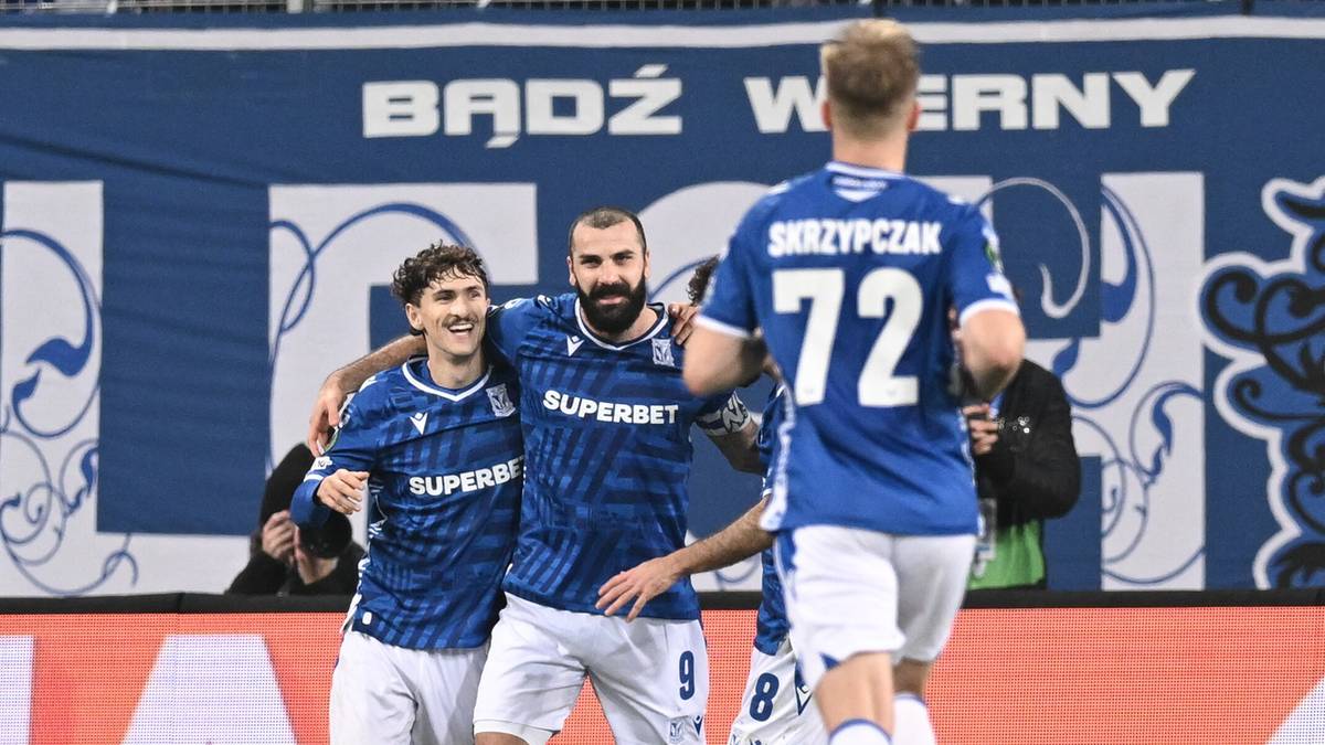 Trzech piłkarzy w niebieskich strojach cieszy się po strzeleniu bramki na tle trybun i baneru "Bądźmy wierni".