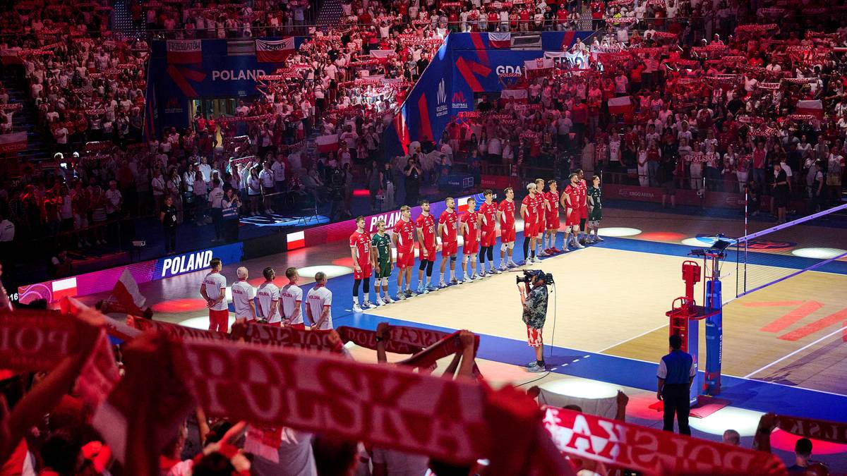 Drużyna siatkarzy Polski ustawiona na boisku w pełnym składzie, z widownią kibiców na trybunach.