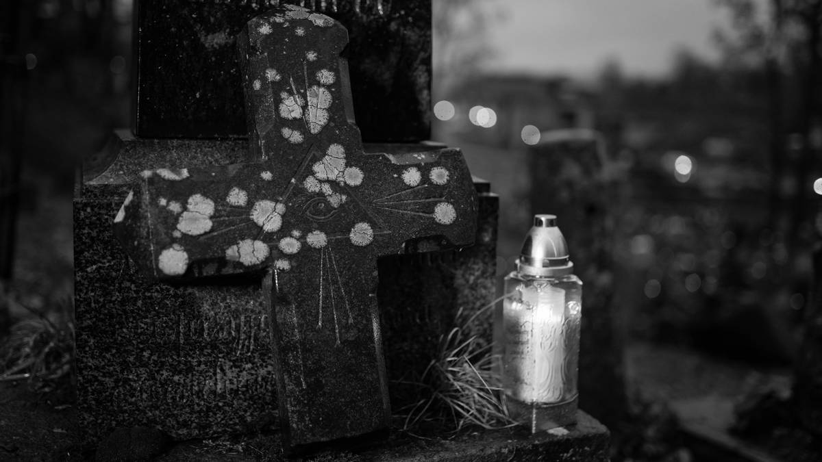 Kamienny krzyż nagrobny z ozdobnymi plamami i płonącym zniczem obok, na tle cmentarza o zmierzchu.