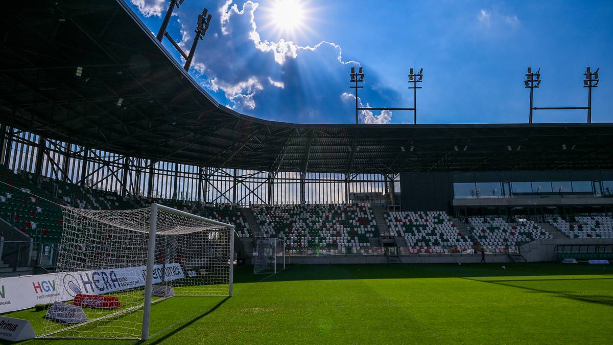 Widok na pusty stadion piłkarski z murawą, bramką i trybunami pod błękitnym niebem z promieniami słońca przebijającymi się przez chmury.