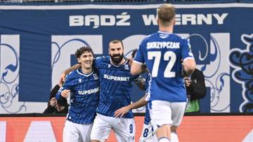 Trzech piłkarzy w niebieskich strojach cieszy się po strzeleniu bramki na tle trybun i baneru "Bądźmy wierni".