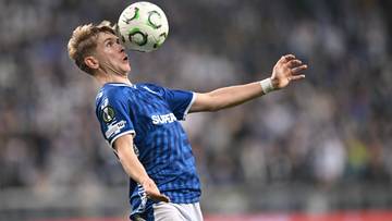 Piłkarz w niebieskiej koszulce z białym kołnierzykiem i białymi spodenkami zagrywa głową piłkę nożną podczas meczu.