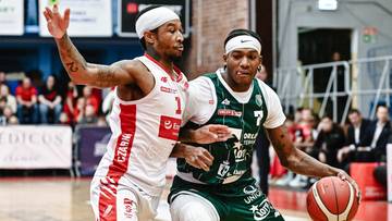 Dw&oacute;ch koszykarzy w akcji podczas meczu. Jeden zawodnik w białej koszulce z czerwonymi elementami, drugi w zielonej z białymi detalami. Obaj mają na głowach opaski.