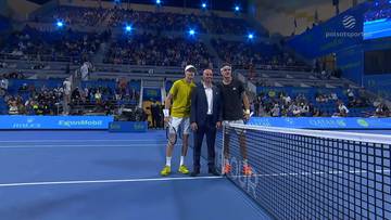 Dw&oacute;ch tenisist&oacute;w i sędzia stoją na korcie tenisowym przed trybunami pełnymi kibic&oacute;w.